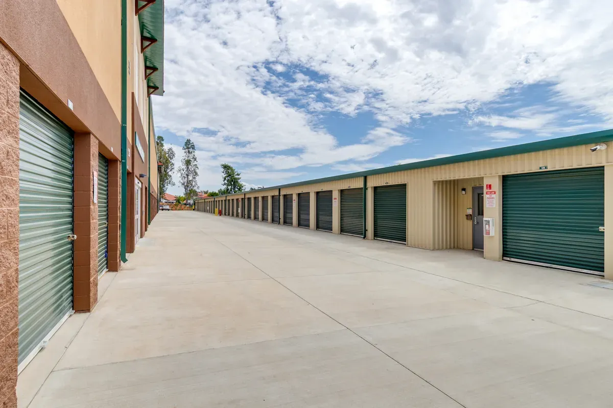 Wide paved drive aisle lined with drive-up self-storage units with dark green roll-up doors under a blue sky.