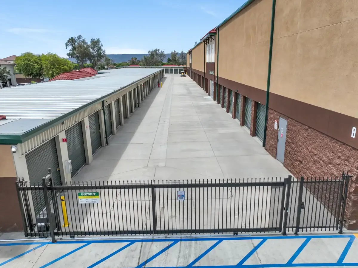 A wide, gated concrete drive aisle lined with self-storage units featuring roll-up doors and a two-story building.