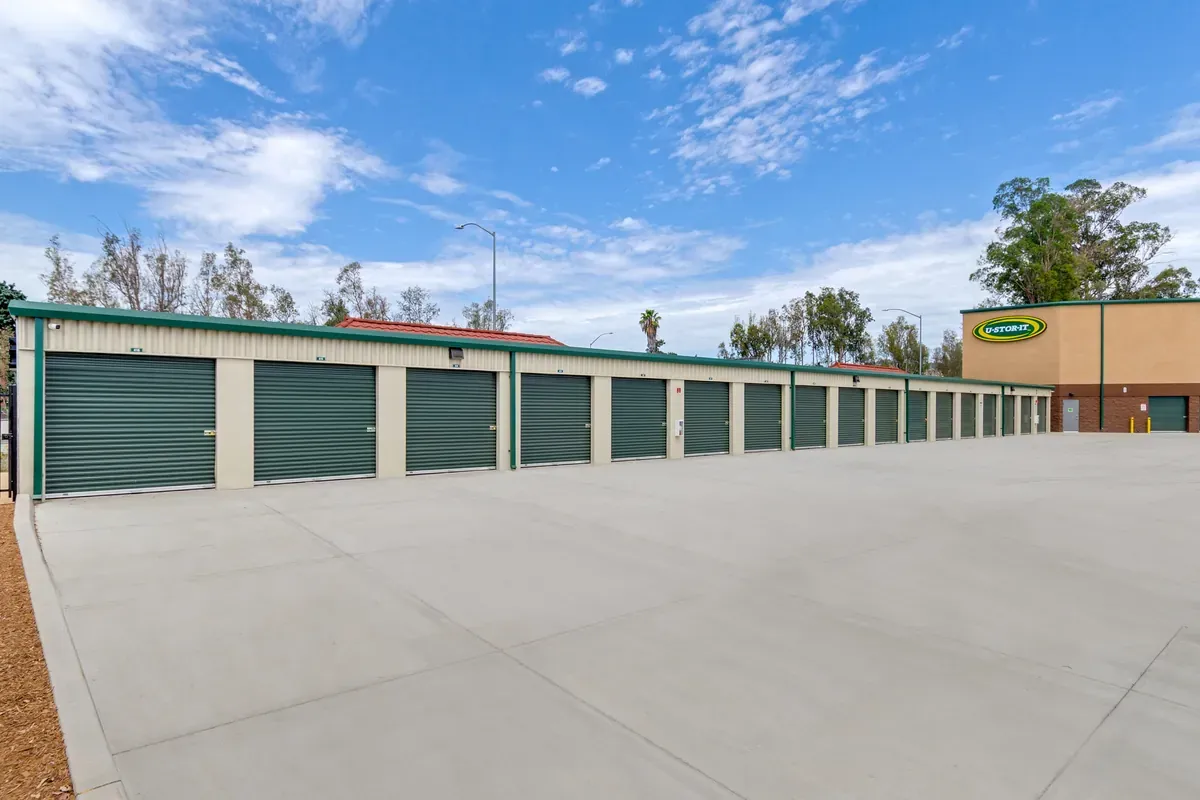 Outdoor drive-up self-storage units with dark green roll-up doors and a wide concrete driveway.